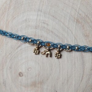 Puede incluir: Un collar choker de macram&eacute; azul con cuentas y abalorios dorados. Los abalorios incluyen dos formas de flor y un peque&ntilde;o elefante. Tambi&eacute;n hay una peque&ntilde;a cuenta redonda de color &aacute;mbar.