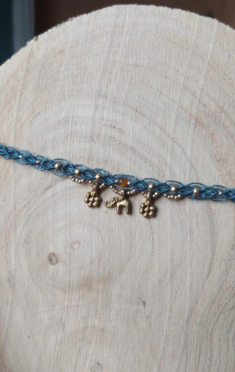 Puede incluir: Un collar choker de macram&eacute; azul con cuentas y abalorios dorados. Los abalorios incluyen dos formas de flor y un peque&ntilde;o elefante. Tambi&eacute;n hay una peque&ntilde;a cuenta redonda de color &aacute;mbar.