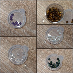 Puede incluir: Cinco peque&ntilde;os recipientes de pl&aacute;stico transparente contienen cada uno un tipo diferente de cuentas de piedras preciosas. Las cuentas son moradas, marrones, transparentes, blancas y verde oscuro. Los recipientes est&aacute;n dispuestos sobre una superficie de madera marr&oacute;n claro.