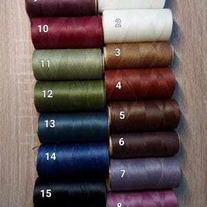 Puede incluir: Una colecci&oacute;n de quince carretes de hilo en varios colores, incluyendo blanco, crema, marr&oacute;n, verde, azul, morado y negro. Cada carrete est&aacute; numerado del 1 al 15. Los hilos est&aacute;n dispuestos sobre una superficie de madera.