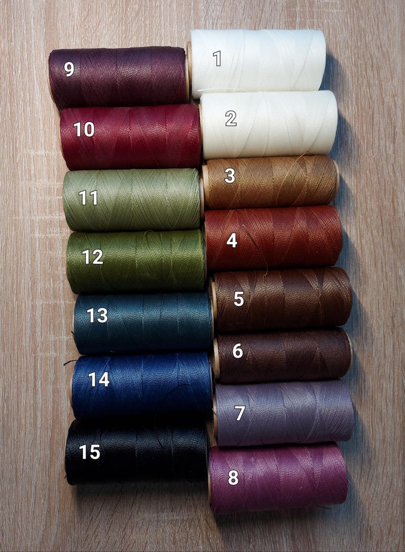 Puede incluir: Una colecci&oacute;n de quince carretes de hilo en varios colores, incluyendo blanco, crema, marr&oacute;n, verde, azul, morado y negro. Cada carrete est&aacute; numerado del 1 al 15. Los hilos est&aacute;n dispuestos sobre una superficie de madera.