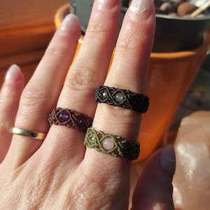 Könnte beinhalten: Nahaufnahme von mehreren Makramee-Ringen in verschiedenen Farben, darunter Braun, Schwarz und Grün. Jeder Ring hat einen zentralen Edelstein in Lila-, Rosa- und Klartönen. Ein einfacher Goldring ist ebenfalls sichtbar.