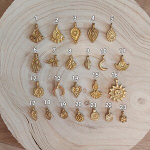 Puede incluir: Una colecci&oacute;n de veintitr&eacute;s colgantes dorados con diversos dise&ntilde;os, incluyendo gotas, lunas, soles y formas geom&eacute;tricas. Los colgantes se exhiben sobre una superficie de madera marr&oacute;n claro, mostrando sus intrincados detalles y texturas. Cada colgante est&aacute; numerado.