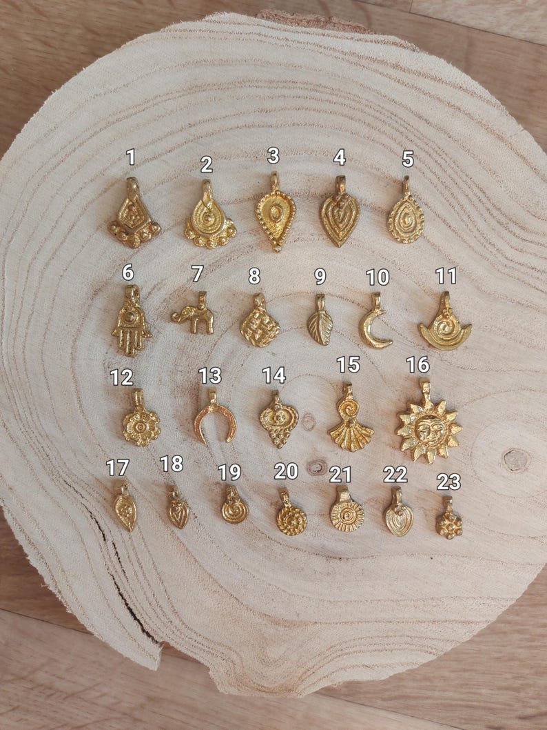 Puede incluir: Una colecci&oacute;n de veintitr&eacute;s colgantes dorados con diversos dise&ntilde;os, incluyendo gotas, lunas, soles y formas geom&eacute;tricas. Los colgantes se exhiben sobre una superficie de madera marr&oacute;n claro, mostrando sus intrincados detalles y texturas. Cada colgante est&aacute; numerado.