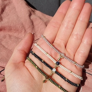 Könnte beinhalten: Fünf handgefertigte Makramee-Armbänder in verschiedenen Farben, darunter Olivgrün, Schwarz, Weiß und Grau. Jedes Armband hat kleine goldene Perlen und einen dekorativen Anhänger mit einem Edelstein. Die Armbänder werden auf einer Hand präsentiert.