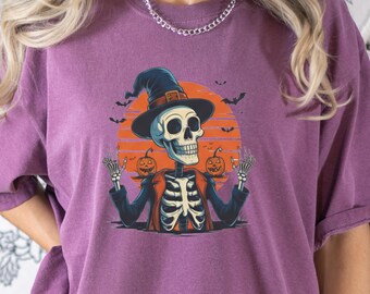 May include: A plum-coloured t-shirt featuring a graphic of a skeleton wearing a black witch hat and an orange jacket. The skeleton is positioned in front of a large orange moon with two jack-o'-lanterns and several small black bats flying around.
