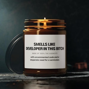 Könnte beinhalten: Ein brennendes braunes Glas-Kerzenglas mit schwarzem Deckel. Das Etikett lautet "SMELLS LIKE DEVELOPER IN THIS BITCH" mit Text über Code und Semikolons. Die Kerze steht auf einer dunklen Oberfläche.