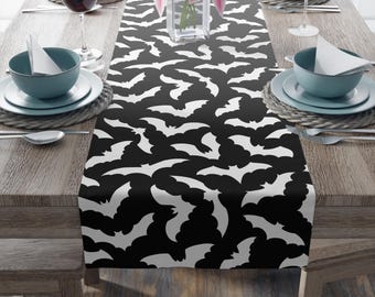 May include: A black and white table runner featuring a repeating pattern of stylized bats. The bats are white against a black background. The runner is placed on a wooden table set for a meal, with plates, bowls, and wine glasses.
