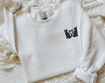 Peut inclure: Un sweat-shirt blanc avec une silhouette noire de deux chats formant un cœur sur le devant.