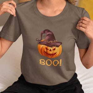 Puede incluir: Una camiseta gris con un diseño de Halloween. El diseño incluye una calabaza naranja con un sombrero de bruja morado oscuro. La palabra "BOO!" está impresa en naranja debajo de la calabaza.