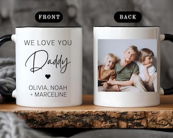 Puede incluir: Taza de cerámica blanca con asa y borde negros. La parte delantera de la taza dice "WE LOVE YOU Daddy" con un corazón y los nombres Olivia, Noah y Marceline. La parte trasera presenta una foto de tres niños.