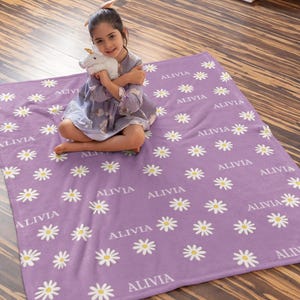 May include: A lavender blanket with a daisy pattern and the name "ALIVIA" repeated throughout. The daisies have white petals and yellow centers. The blanket is made of a soft material.
