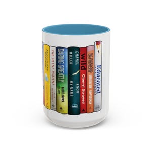 May include: White ceramic mug with a blue rim, featuring a design of a stack of colorful books with titles like "Educated", "Wild", "Daring Greatly", and "The Silent Patient".