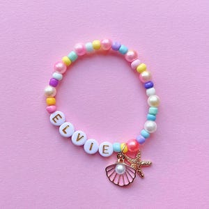 May include: A pastel-coloured beaded bracelet with the name "ELVIE" spelled out in white letter beads. The bracelet has a gold seashell and starfish charm. The beads are in shades of pink, yellow, blue, and purple.