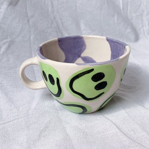 Könnte beinhalten: Eine weiße Keramiktasse mit einem lila und grünen Design. Die Tasse hat einen Henkel und ist mit grünen Smiley-Gesichtern verziert.