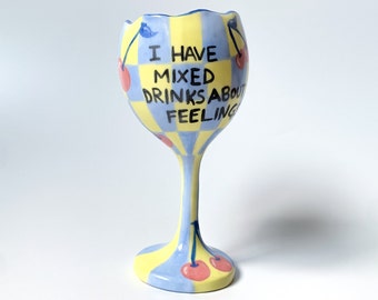 May include: A blue and yellow ceramic wine glass with a checkered pattern and cherries painted on it. The glass has the text "I have mixed drinks about feeling" written on it.