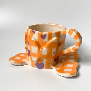 Könnte beinhalten: Eine Teetasse und ein Unterteller aus Keramik mit einem Gingham-Muster in Orange und Weiß. Die Tasse hat einen Henkel und ist mit lila Herzen und Schmetterlingen verziert.