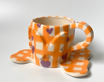 May include: A ceramic teacup and saucer with a gingham pattern in orange and white. The cup has a handle and is decorated with purple hearts and butterflies.