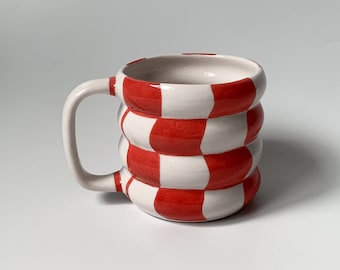 May include: A white ceramic mug with a red and white checkered pattern. The mug has a rounded shape and a handle.