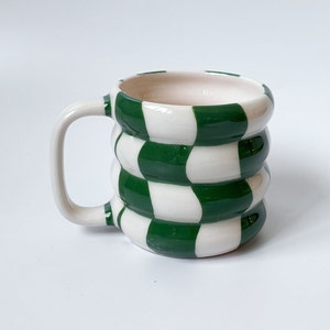 Könnte beinhalten: Eine weiße Keramiktasse mit einem grün-weiß karierten Muster. Die Tasse hat eine abgerundete Form und einen Henkel.