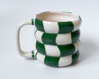 Puede incluir: Una taza de cerámica blanca con un patrón de cuadros verde y blanco. La taza tiene una forma redondeada y un asa.