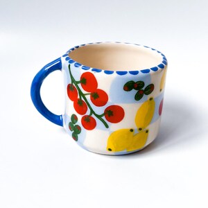 Puede incluir: Una taza de cerámica blanca con asa azul y un patrón a cuadros. La taza está decorada con tomates rojos, limones amarillos y hojas verdes.