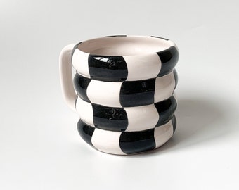 Puede incluir: Una taza de cerámica blanca con un patrón de cuadros negros y blancos. La taza tiene una forma redondeada y un asa.