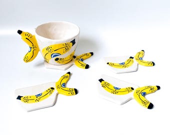 Può includere: Un set di tazze da caffè e sottobicchieri in ceramica bianca. La tazza e i sottobicchieri sono decorati con disegni di bucce di banana gialle con accenti neri e blu. Il set è progettato per assomigliare a un gruppo di banane.