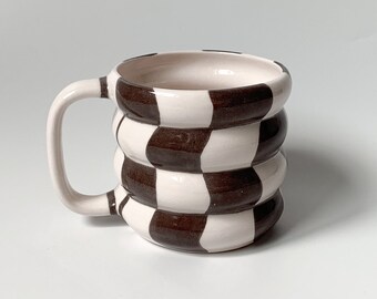Puede incluir: Una taza de cerámica blanca con un patrón de cuadros marrones y blancos. La taza tiene una forma redondeada y un asa.
