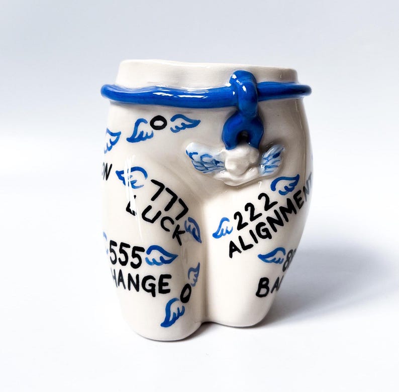 May include: White ceramic mug shaped like a pair of pants with blue trim and wings. The mug has the numbers 777, 555, and 222 with the words "LUCK", "CHANGE", and "ALIGNMENT" written on it.