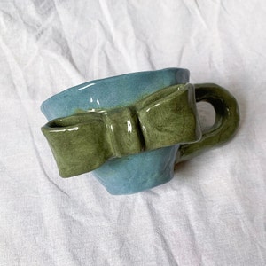 Może przedstawiać: Ręcznie wykonany ceramiczny kubek z jasnoniebieskim korpusem i zieloną kokardą. Kubek ma zaokrąglony uchwyt i lekko nieregularny brzeg, co sugeruje jakość rękodzieła. Tło jest białe.