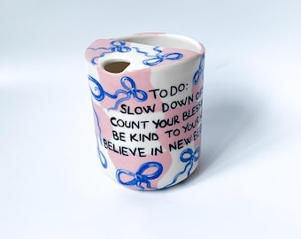 Puede incluir: Una taza de cerámica blanca con un diseño rosa y azul. La taza tiene una cita que dice: "TODO: SLOW DOWN OF COUNT YOUR BLESS BE KIND TO YOUR BELIEVE IN NEW BE".