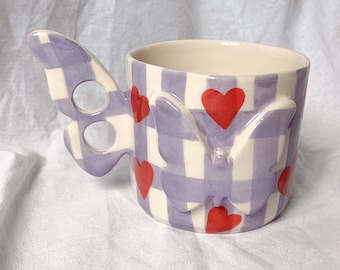 May include: A ceramic mug with a butterfly wing handle and butterfly accents. The mug features a lavender and white checkered pattern with red heart accents. The mug is a unique and decorative item.