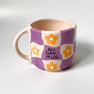 Może przedstawiać: Kubek w kratkę w kolorze fioletowym i białym z żółtymi kwiatami i napisem "ALL DAY MUG" w kolorze czarnym.