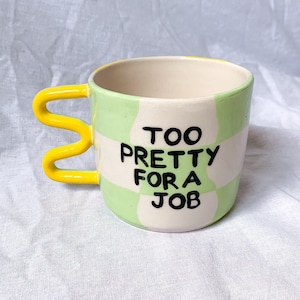 Puede incluir: Una taza de cerámica con interior crema, asa amarilla ondulada y exterior a cuadros verdes y blancos. La taza tiene la frase "TOO PRETTY FOR A JOB" en letras negras.