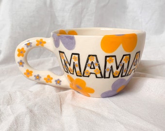 May include: White ceramic mug with a floral design in shades of yellow and purple. The word "MAMA" is written in black outlined letters with a floral pattern inside the letters.