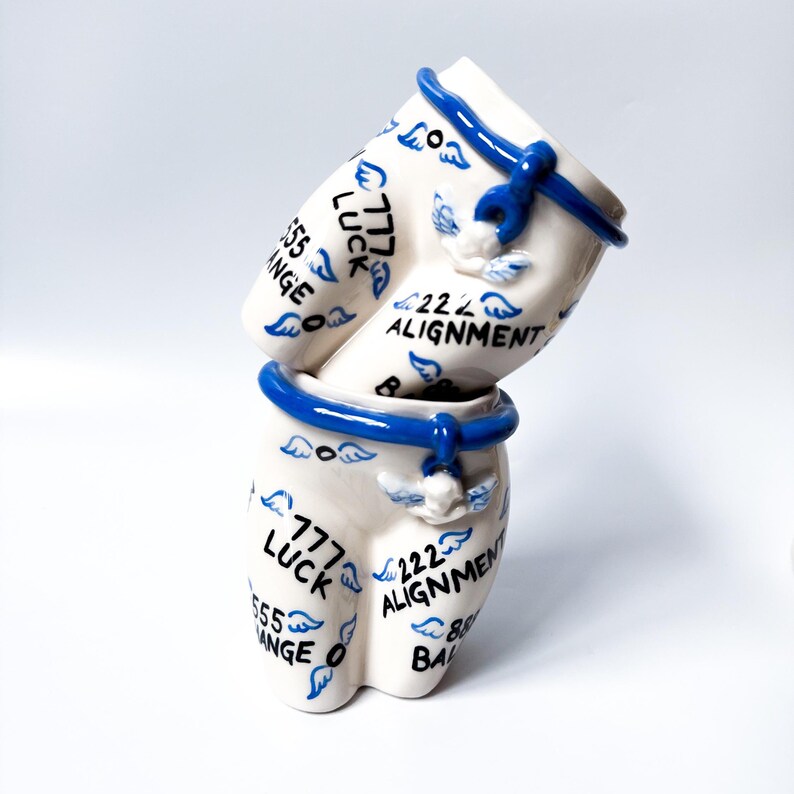 May include: Two white ceramic cups stacked on top of each other. The cups are decorated with blue and white wings and the text "777 Luck", "222 Alignment", "555 Change", and "888 Balance".