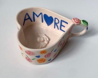 May include: A heart-shaped ceramic dish with a blue "AMORE" inscription and a blue heart. The dish is decorated with pink and yellow flowers and has a handle with a pink flower.