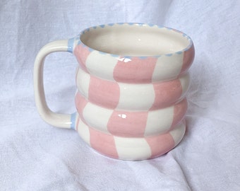 May include: A ceramic mug with a unique design. The mug features a pink and white checkered pattern on the body, a white handle, and a light blue rim. The mug is designed with a rounded, stacked shape.