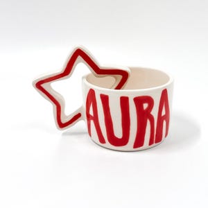 May include: A white ceramic mug featuring the word AURA painted in bold red letters on the front. The mug has a unique handle shaped like a five-pointed star with a red outline, providing a distinct visual element to the cylindrical vessel.