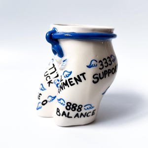May include: White ceramic cup with a blue rim and a blue band around the top. The cup is decorated with blue wings and black text that reads "777 Luck", "Enviornment Support", "333", and "888 Balance".