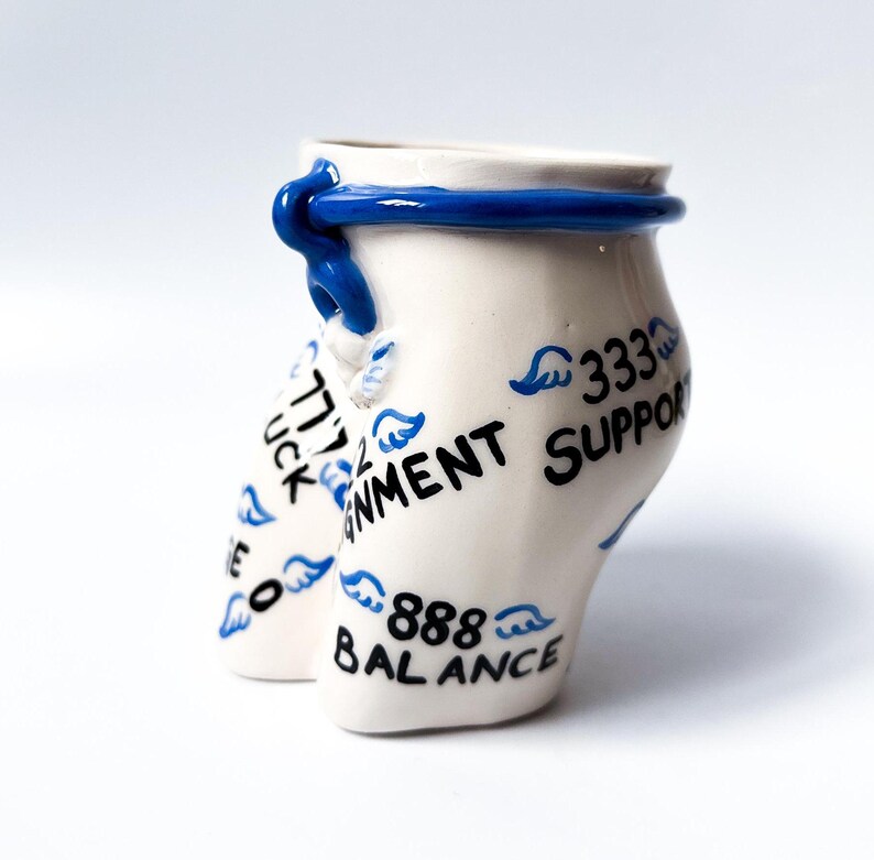 May include: White ceramic cup with a blue rim and a blue band around the top. The cup is decorated with blue wings and black text that reads "777 Luck", "Enviornment Support", "333", and "888 Balance".