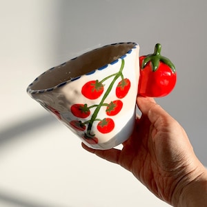 Puede incluir: Una taza de cerámica con diseño de tomate. La taza es blanca con un borde festoneado azul y un asa con forma de tomate rojo. La taza presenta una vid verde con tomates rojos pintados en el lateral.