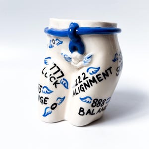 May include: White ceramic figurine shaped like a human body with blue painted details. The figurine has various words and numbers written on it, including "777 Lick", "222 Alignment", and "888 Balance".