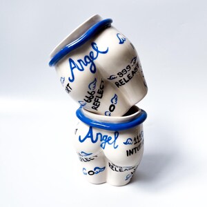 May include: Two white ceramic mugs with blue trim and the word "Angel" written on them. The mugs are decorated with blue wings and the words "Reflect", "Release", "Intuition", and "999".