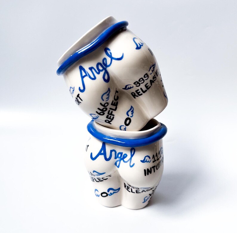 May include: Two white ceramic mugs with blue trim and the word "Angel" written on them. The mugs are decorated with blue wings and the words "Reflect", "Release", "Intuition", and "999".
