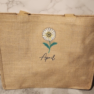 May include: A rectangular burlap tote bag with a daisy embroidered on the front. The daisy has white petals, a yellow center, and a green stem. The word "April" is written below the flower in black script.