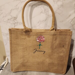 May include: A brown burlap tote bag with a pink flower and the word "January" embroidered on the front.