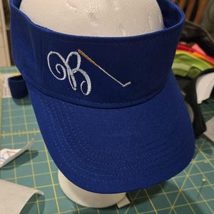 May include: A royal blue visor with a curved brim. The visor features an embroidered white script initial "B" with a golf club design. The visor is displayed on a white mannequin head.