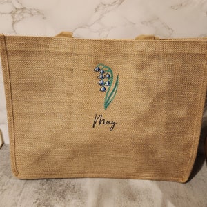May include: A brown burlap tote bag with a green and blue lily of the valley design and the word "May" embroidered on it.